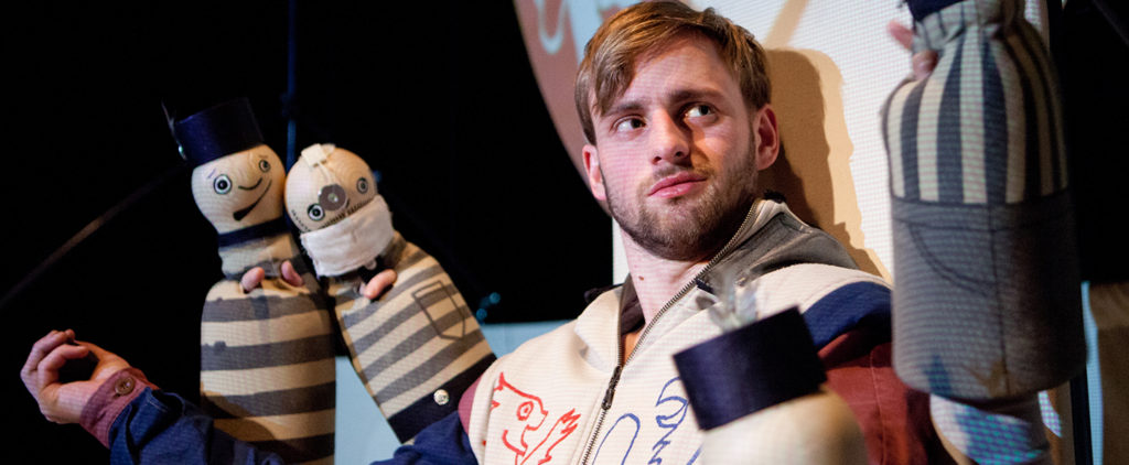 Actor playing King Charles seated on a throne with two hand puppets beside him.