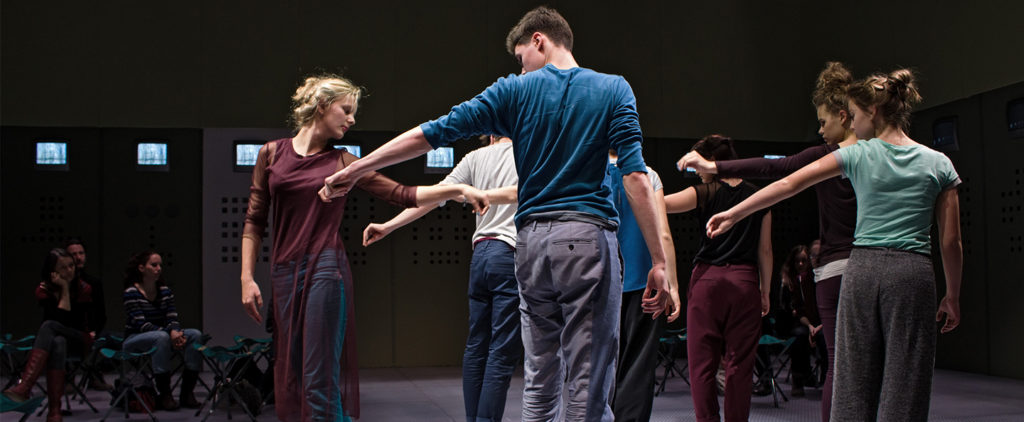 Performers stand in a circle, each with their left arms extended, gazing towards their hands.